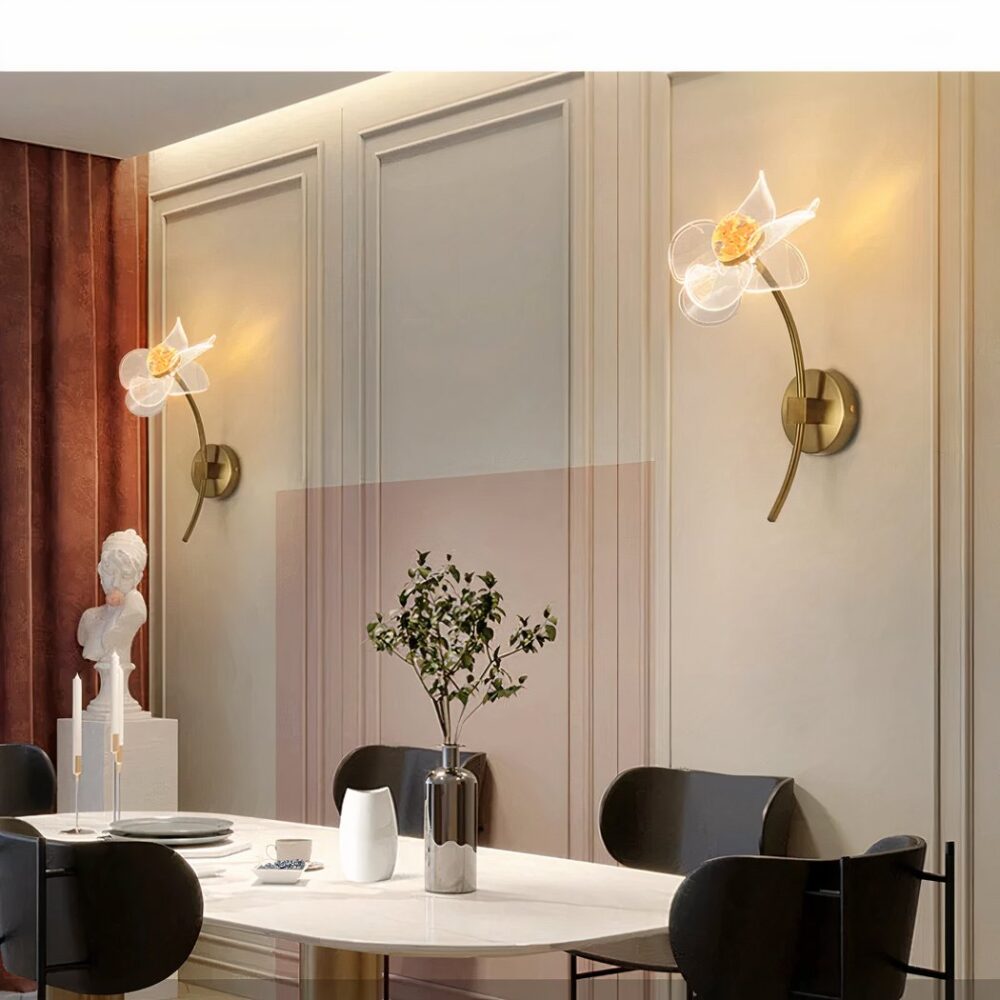 Großer Raum mit weißem Tisch und vier schwarzen Stühlen darum herum. Eine Vase mit Laub, Kerzenhalter und ein weißes Glas stehen auf dem Tisch. Links ein langer brauner Vorhang und eine weiße Statue davor. An der gegenüberliegenden Wand hängen zwei Wandlampen in Form von Blumen mit Stielen, die eingeschaltet sind.