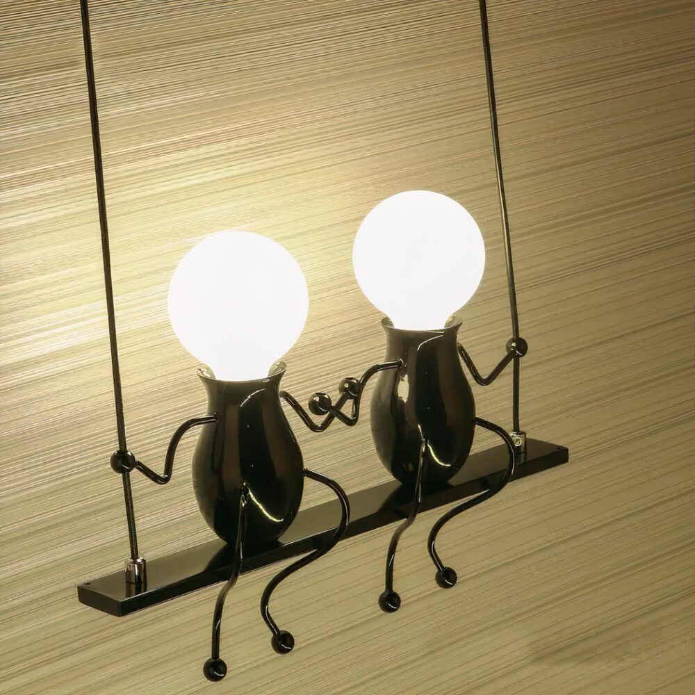 Wandleuchte in Form einer Schaukel mit 2 darauf sitzenden Männchen, deren Kopf die Glühbirne ist. Sie sind eingeschaltet. Hellbrauner Hintergrund dahinter.