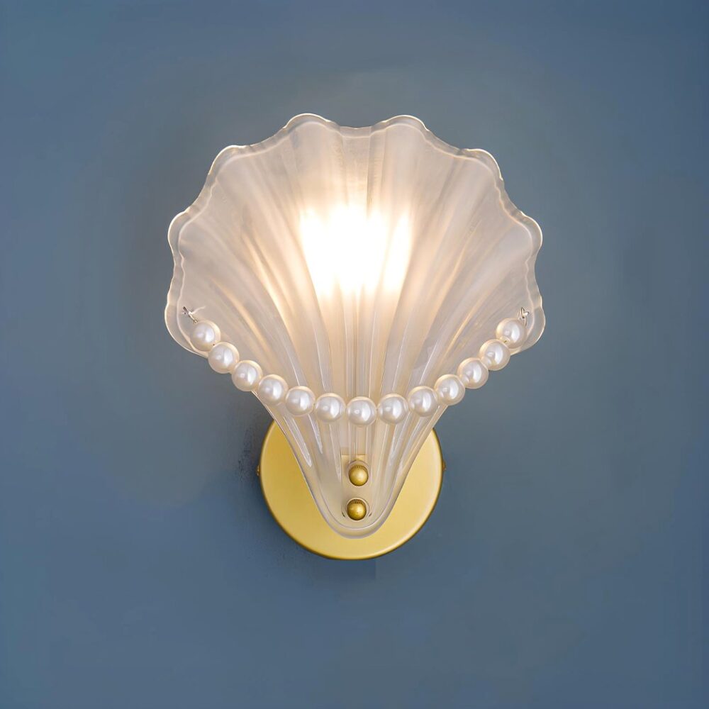 Muschelförmige Wandleuchte aus geschliffenem Glas mit weißen Perlen am Rand der Muschel. Die goldene runde Halterung ist an einer blauen Wand befestigt. Die Glühbirne ist eingeschaltet.