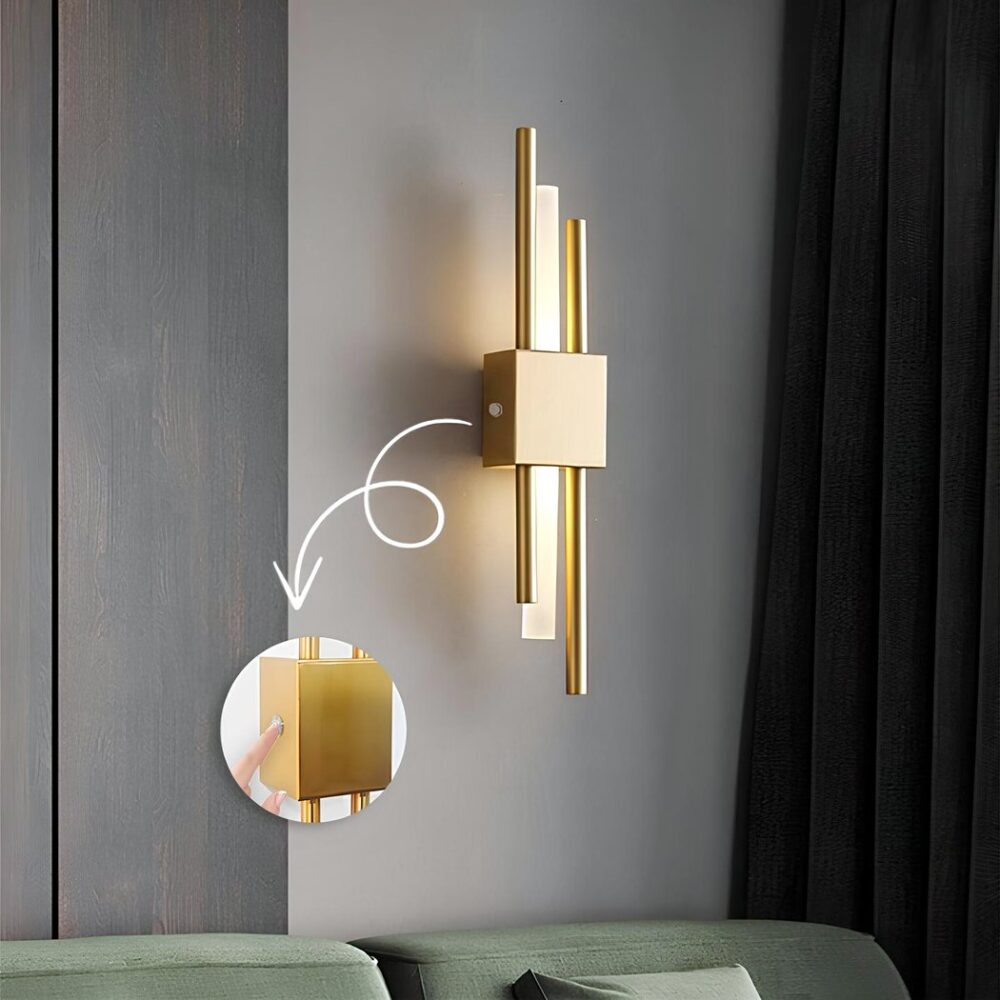 Wandleuchte mit drei Röhren: eine helle Röhre zwischen zwei goldenen Röhren, eine höhere und eine niedrigere, die an einer grauen Wand mit einem dunkelgrauen Vorhang rechts befestigt ist. Ein grünes Sofa unter der Wandleuchte. Eine runde Vignette zeigt den Lichtschalter in Großaufnahme. Ein weißer Pfeil von der Wandleuchte zu dieser Vignette.