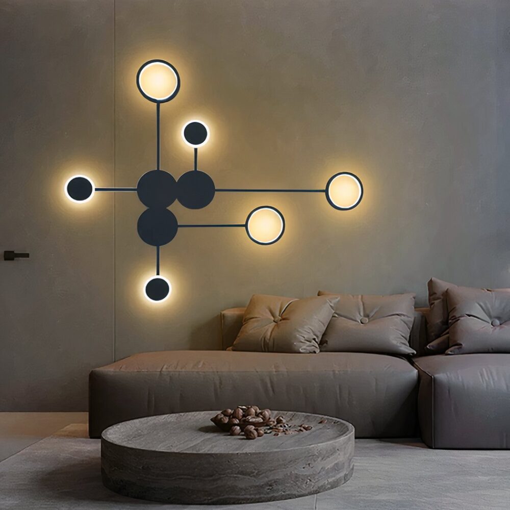 Schwarze Wandleuchte aus Kreisen, die durch Striche miteinander verbunden sind. 3 schwarze Kreise in der Mitte und dann 6 Kreise rundherum leuchtend. Befestigt an einer braunen Wand über einem braunen Sofa mit passenden Kissen. runder brauner Couchtisch und beiger Teppich darunter.