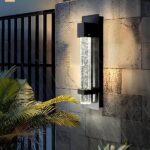 Außenwandleuchte in Form einer Blasenröhre in einer schwarzen Aluminiumstruktur, die an einer Steinmauer befestigt ist. Links ein großes schwarzes Tor, ein Teil einer Palme vor der Wand und fallender Regen.