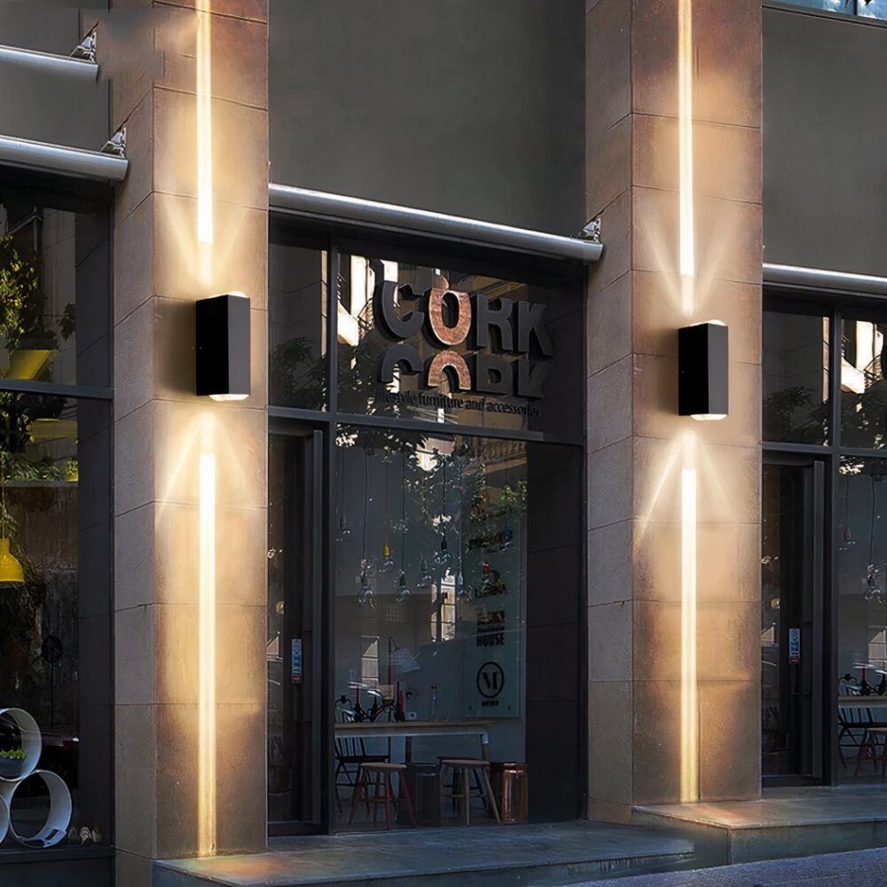 Ladenfront mit Schaufenstern, die von Betonpfeilern eingerahmt sind, an denen jeweils eine Wandleuchte befestigt ist. Die Wandleuchte ist rechteckig und schwarz und strahlt über und unter ihr warmes Licht aus.