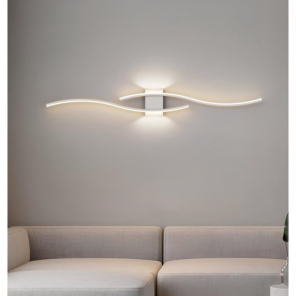 Weiße Wandleuchte in Form von zwei leuchtenden Wellen. Das Licht wird auch oberhalb und unterhalb der rechteckigen Halterung gestreut, die an einer weißen Wand über einem beigen Sofa befestigt ist.