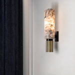 Wandleuchte mit einer Halterung aus schwarzem Metall, die an einer weißen Wand befestigt ist. Der obere Teil der Wandleuchte ist ein Marmorrohr mit einer Glühbirne im Inneren, die das Licht streut. Der untere Teil ist ein graues, geriffeltes Kupferrohr. Ein grau-weiß gestreifter Vorhang auf der linken Seite.