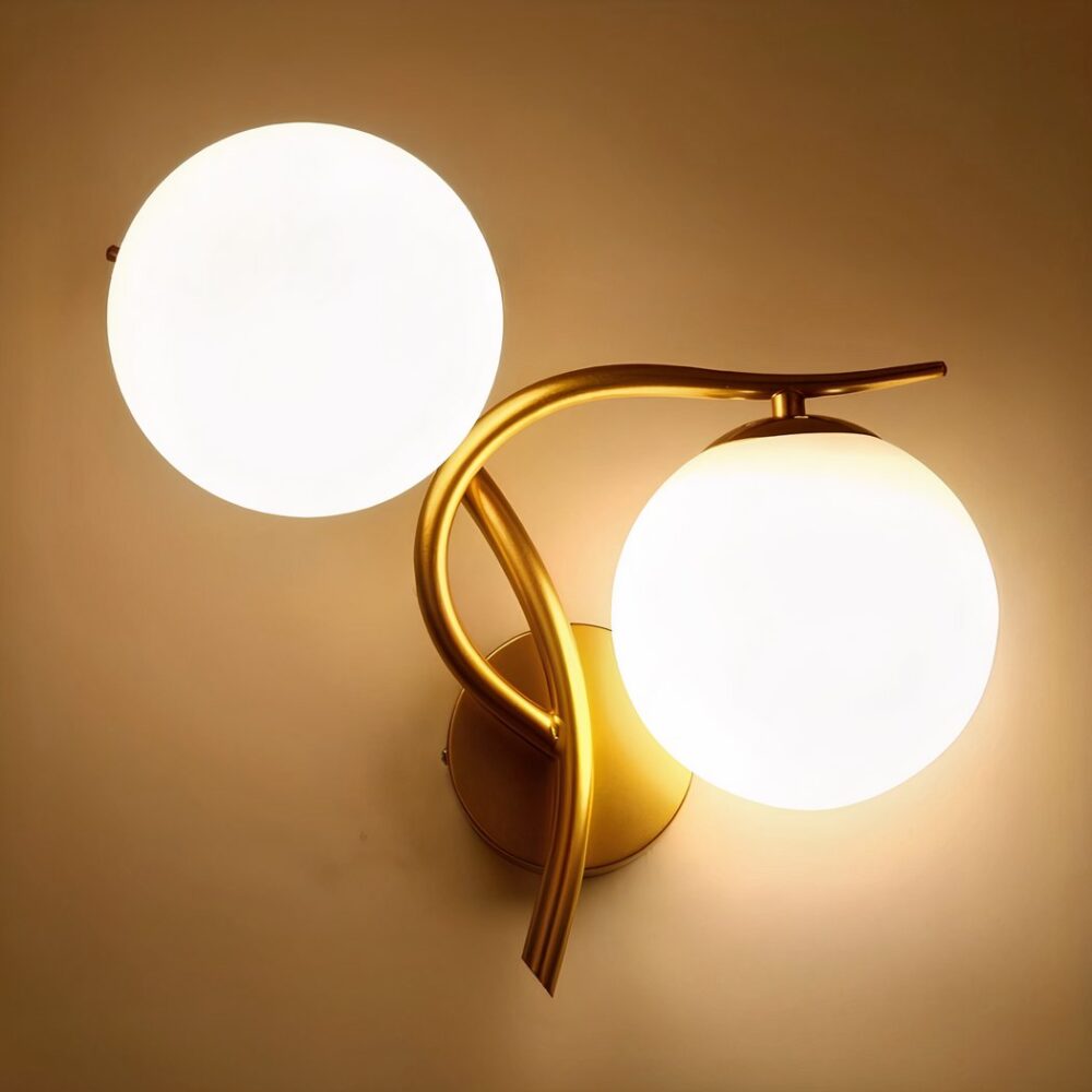 Wandleuchte aus goldfarbenem Metall mit einer Leuchtkugel oberhalb des vertikalen Stabs und der anderen Kugel unterhalb des horizontalen Stabs auf der rechten Seite. Beide Leuchtkugeln sind eingeschaltet. An einer beigen Wand befestigt.