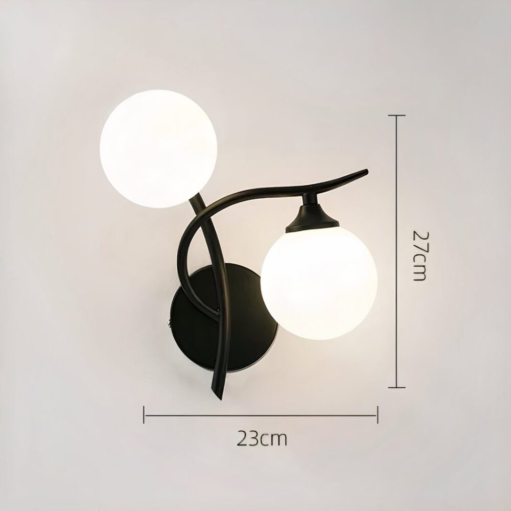 Wandleuchte aus schwarzem Metall mit 2 Leuchtkugeln, eine am vertikalen Stab und eine unter dem horizontalen Stab auf der rechten Seite. Wird an einer grauen Wand befestigt. Die Maße erscheinen links und unten an der Wandleuchte.